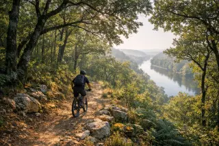 Solitary mountain biker rides through an oak forest trail at dawn with misty light and the Dordogne River visible in the distance.