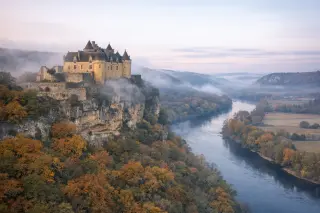 Château de Fénelon at dawn, mist curling around its golden stone walls atop a cliff, surrounded by autumn trees and the Dordogne valley below.